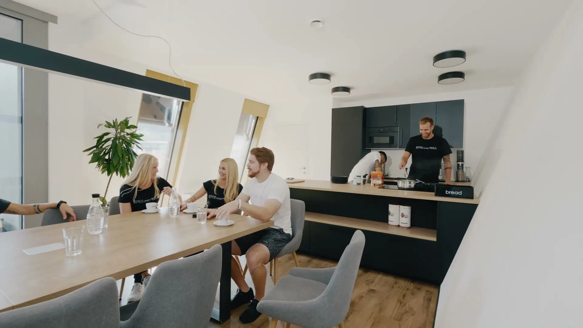 Team von Physio 1.0 am Besprechungstisch im Teamraum, dahinter wird in der Teamküche gekocht